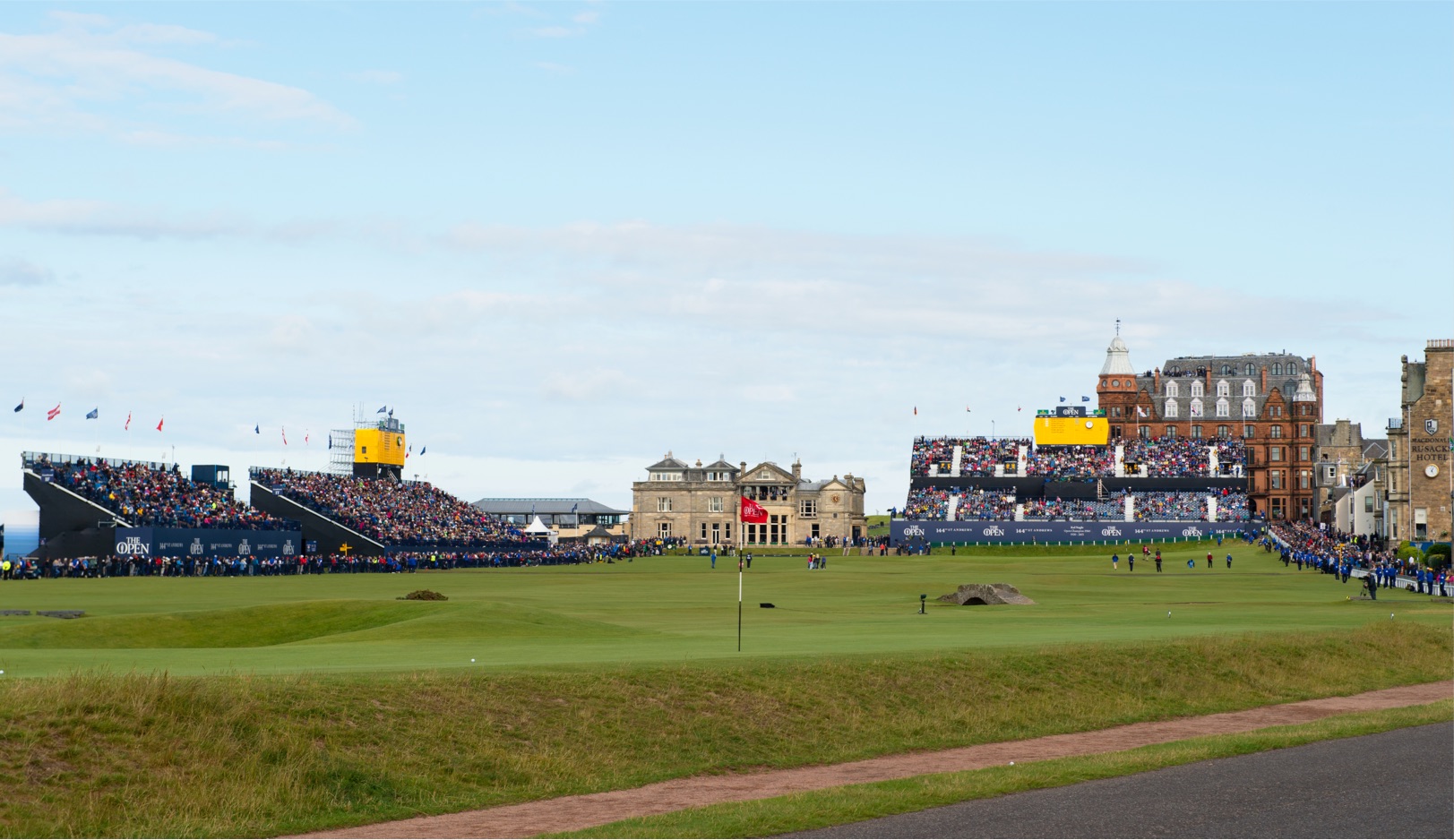 Aerial view of a large golf course with a significant crowd seated in grandstands surrounding the greens. Various structures and buildings are seen nearby, and numerous flags are raised. Golfers and spectators are scattered throughout the grass.