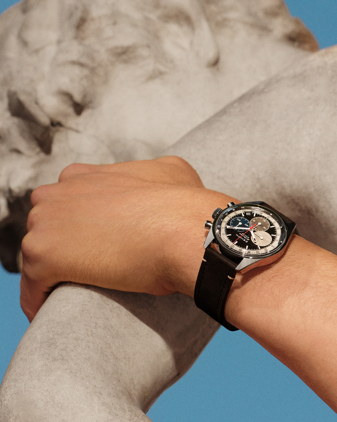 A person wearing a blue sweater with a white trim has their hand in the pocket of black pants. They are wearing a black analog wristwatch with a silver bezel and three subdials on their left wrist.