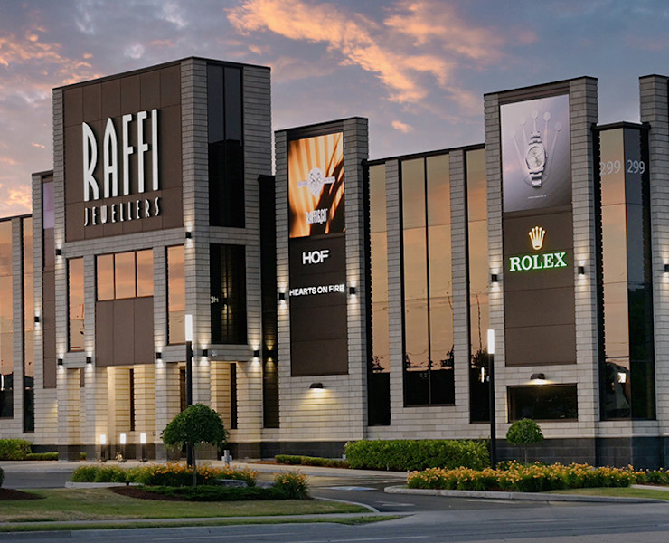 A modern building with multiple lit signs, including "RAFFI JEWELLERS" prominently displayed across several sections. The ROLEX logo also appears on two signs. The structure features large windows and a brick facade, situated on a street corner with cars parked nearby.