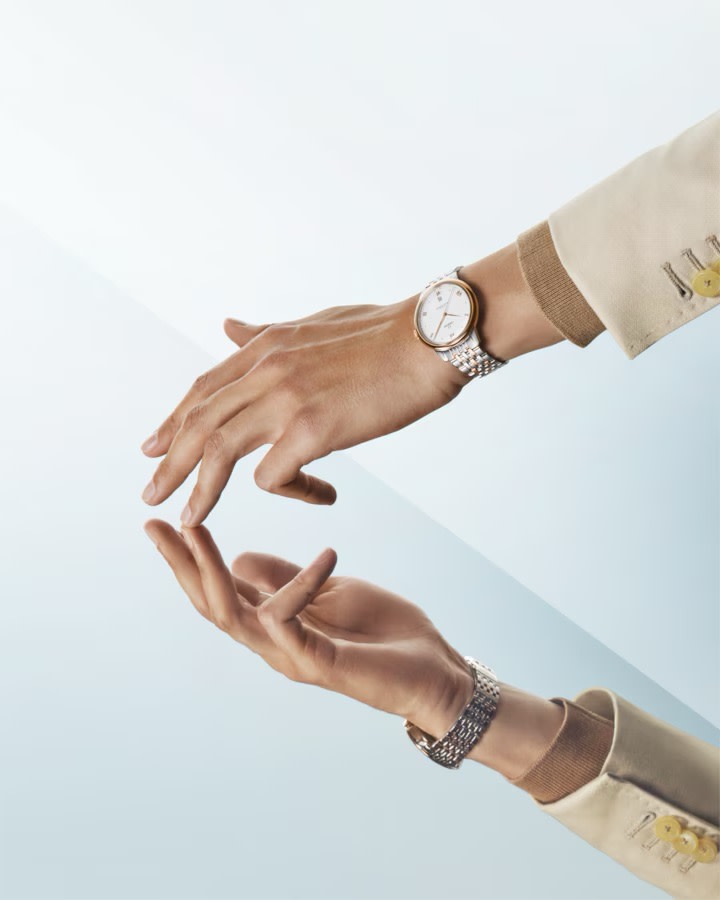Two hands with a minimalist wristwatch with a silver band and white face. The top hand is extended, displaying the watch, while the bottom hand mirrors the top hand's position. Both hands are against a light gradient background.