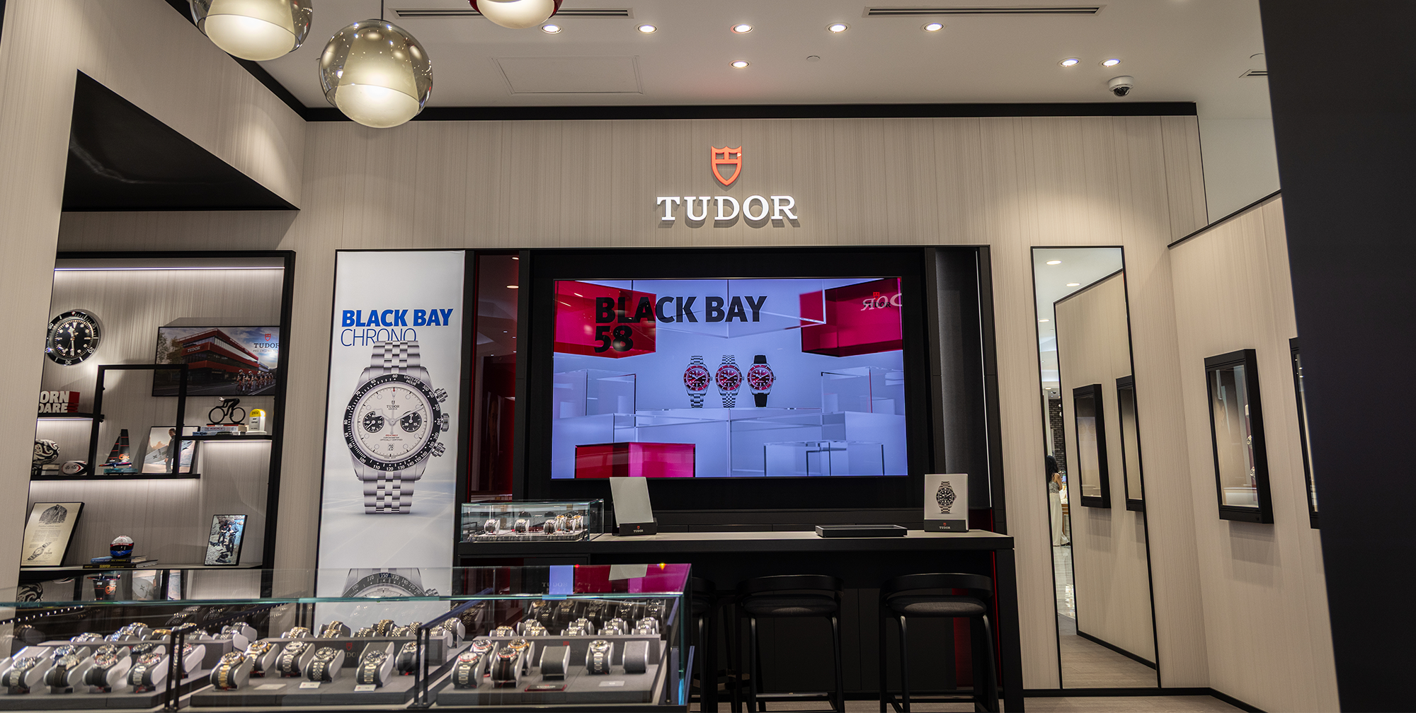 A modern jewelry store interior featuring a TUDOR display with various TUDOR watches. The display includes a large screen showing a close-up of a watch, several framed watches on the wall, and a glass case showcasing watches. A seating area is visible in the background.