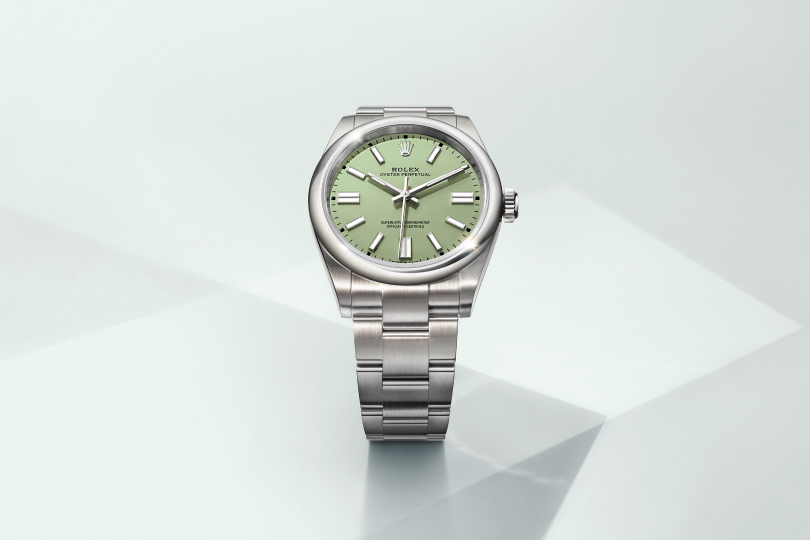 A silver wristwatch with a polished stainless steel strap and a white dial. The watch face features gold hour markers and hands, with the brand name "Rolex" and other details visible on the dial. The background is a soft, geometric light beige.