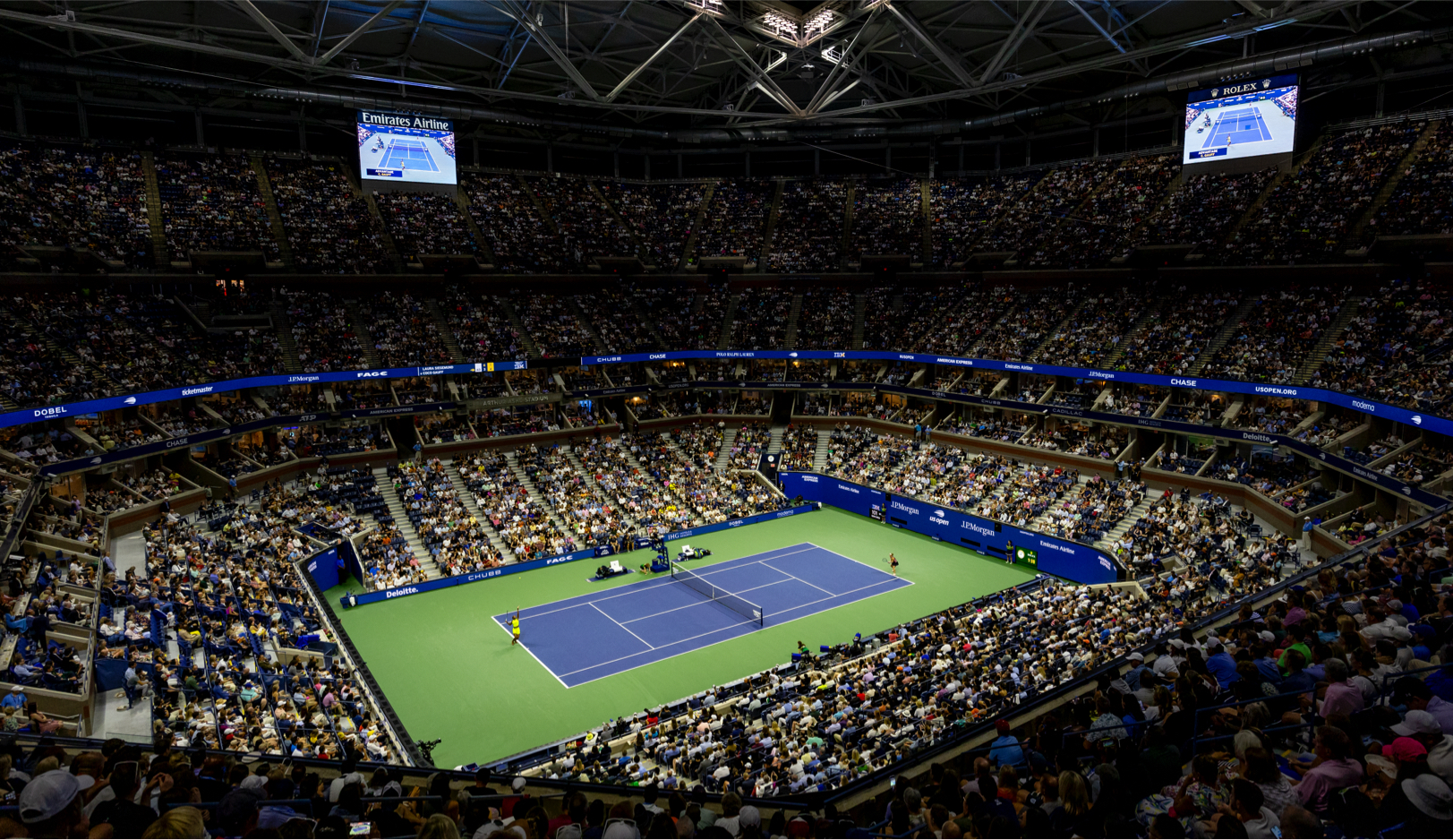 A nighttime view of a packed tennis stadium with thousands of spectators watching a match. The court is illuminated under the closed roof, with players visible in action. The arena screens display match information.