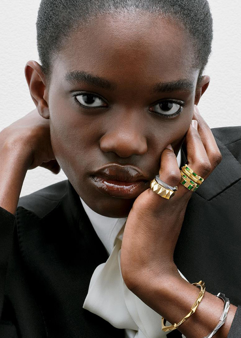 A person with short, dark hair gazes directly at the camera. They rest their face on one hand, fingers adorned with multiple shiny rings, while wearing a black jacket over a white top. The background is plain and light-colored.