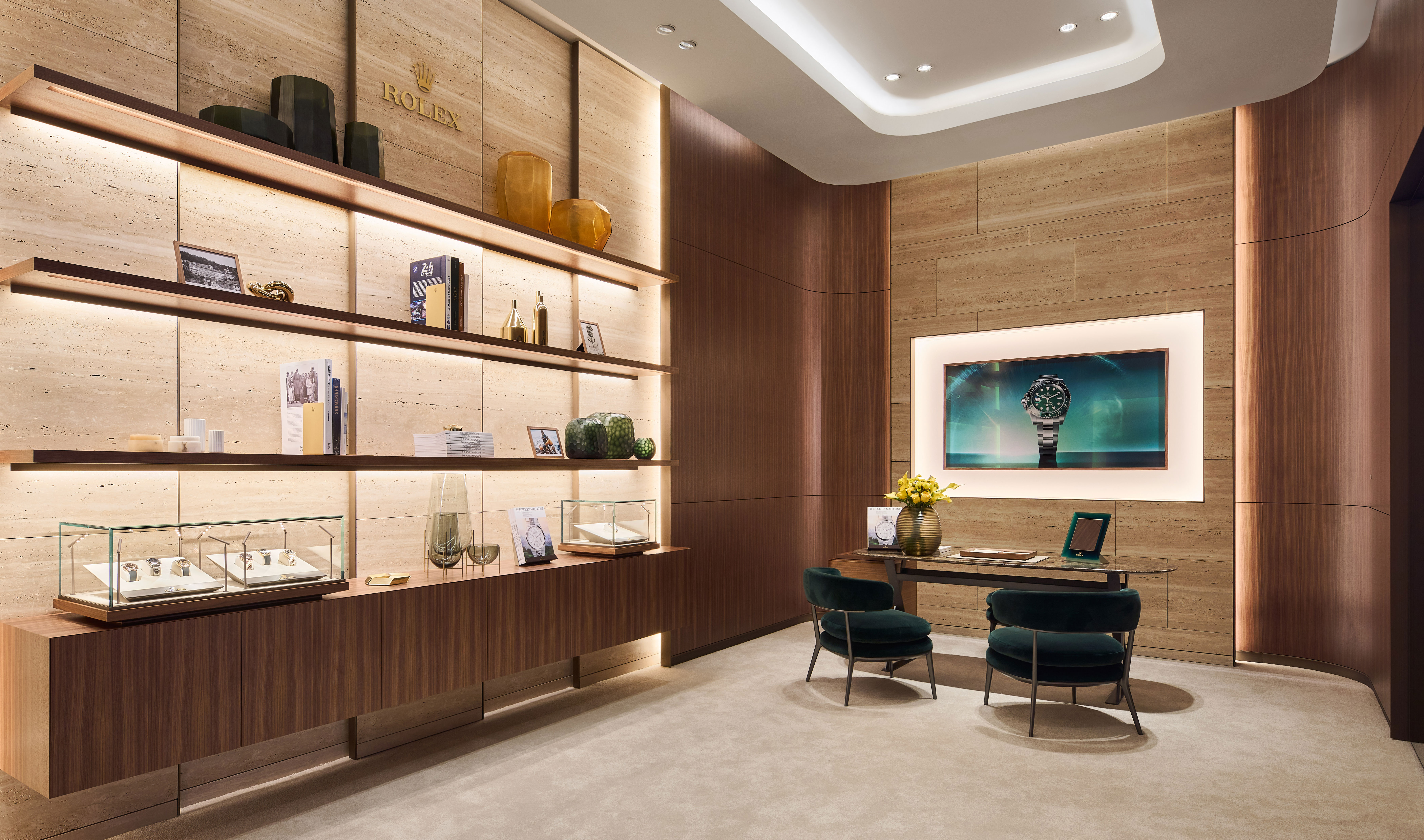 A modern showroom with green cushioned sofas arranged around wooden coffee tables. Wall-mounted display cases showcase Rolex watches, and large windows provide a view of a snowy landscape. The interior has hardwood floors and a minimalist design.