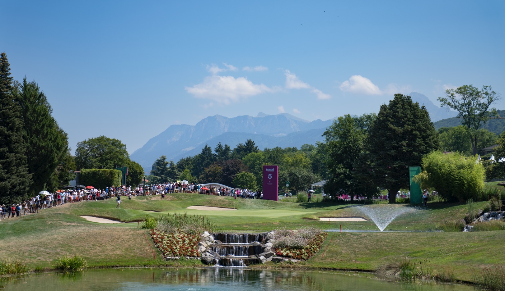 A scenic landscape of a lakeside golf course. The course features well-maintained green lawns, sand traps, and a curving water hazard. Several buildings and a gathering of people are visible, with a serene lake and distant mountains in the background.