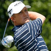 Male golfer at the end of his swing watching the ball fly.  He is wearing a blue golf shirt with horizontal white stripes and a white hat.
