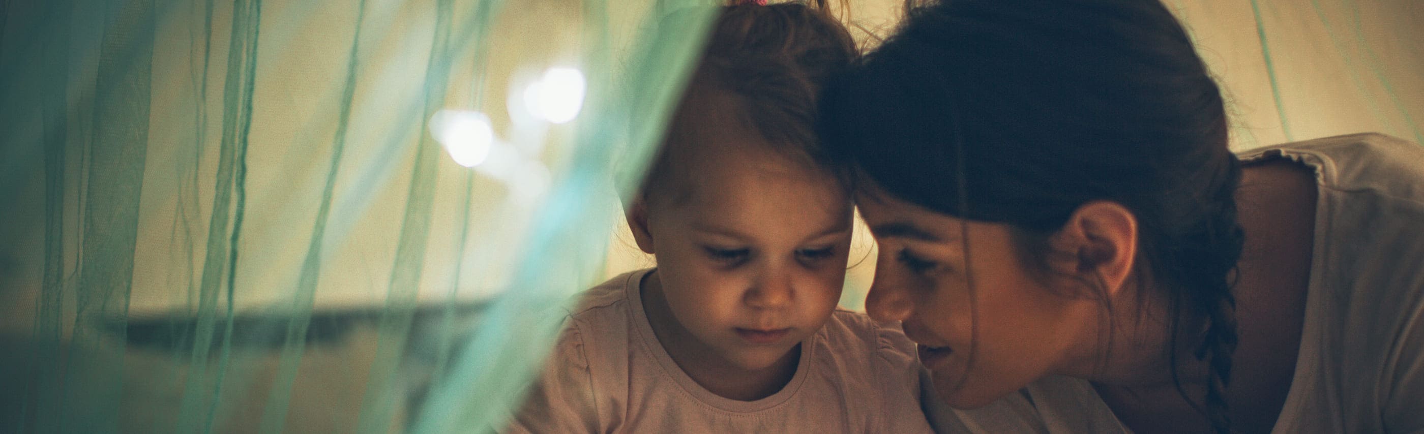 Mother sitting next to her young daughter in her bed with a canopy at night. 