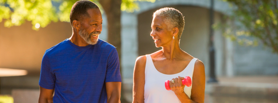 An older man with a blue shirt and a woman with a white tank top with dumbbells in her hands exercising outdoors 