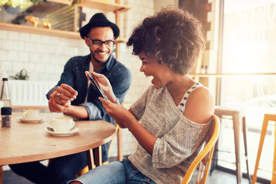 Woman laughing and looking at phone while sitting at a table with a man