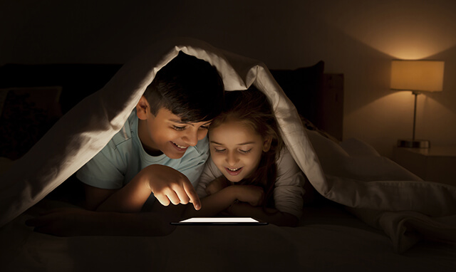 Des enfants installés dans un lit, dans le noir, et lisant sur une tablette 