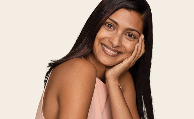 Woman with long dark straight hair holding her face with one hand and smiling