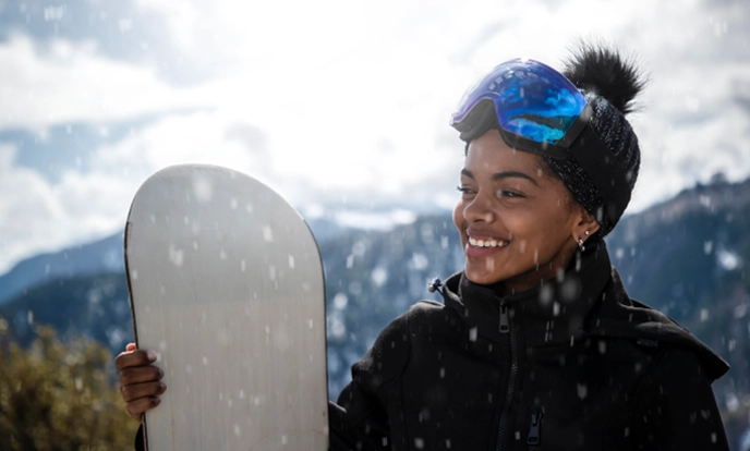 Person holding a snowboard and smiling outdoors in a snowy area.