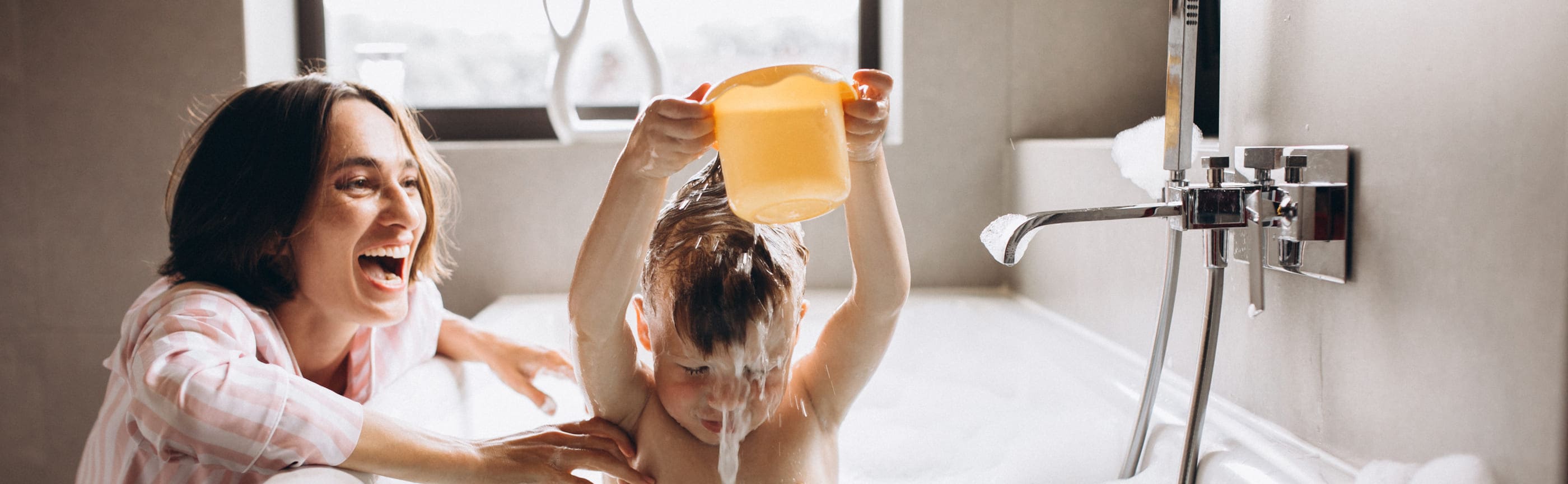 Young mother smiling at her son who is sitting in the tub pouring water on his head.