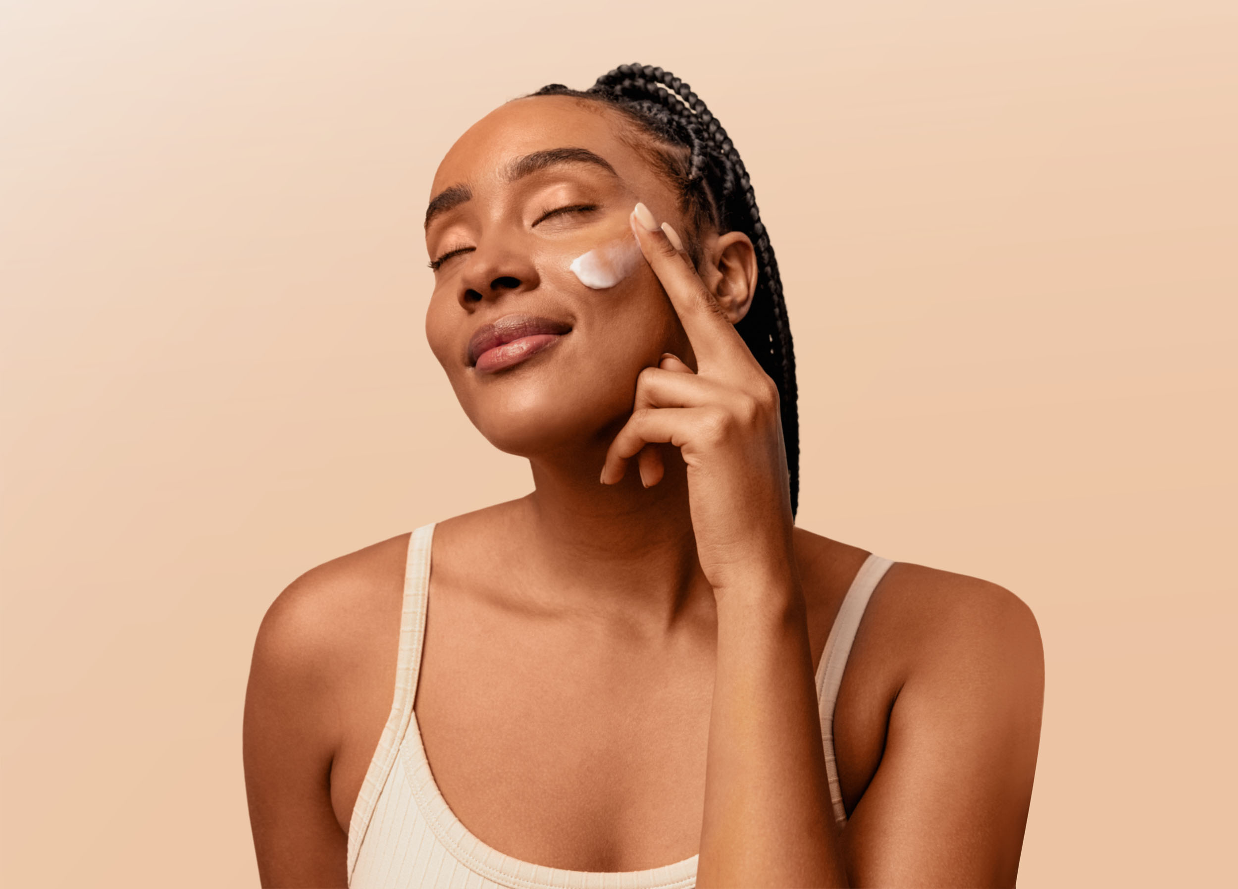 Young woman applying face cream on her face while her eyes closed and smiling