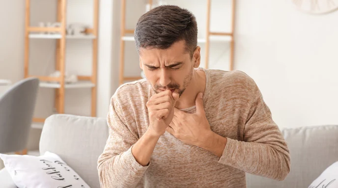 Man sitting on a coach coughing into his fist and holding his chest.