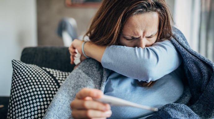 Sick woman sitting and covering her mouth with her left arm while holding a thermometer in her right hand.