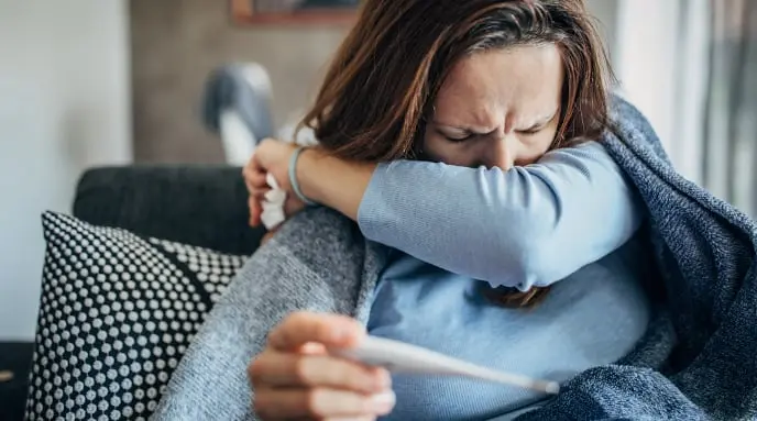 Sick woman sitting and covering her mouth with her left arm while holding a thermometer in her right hand.
