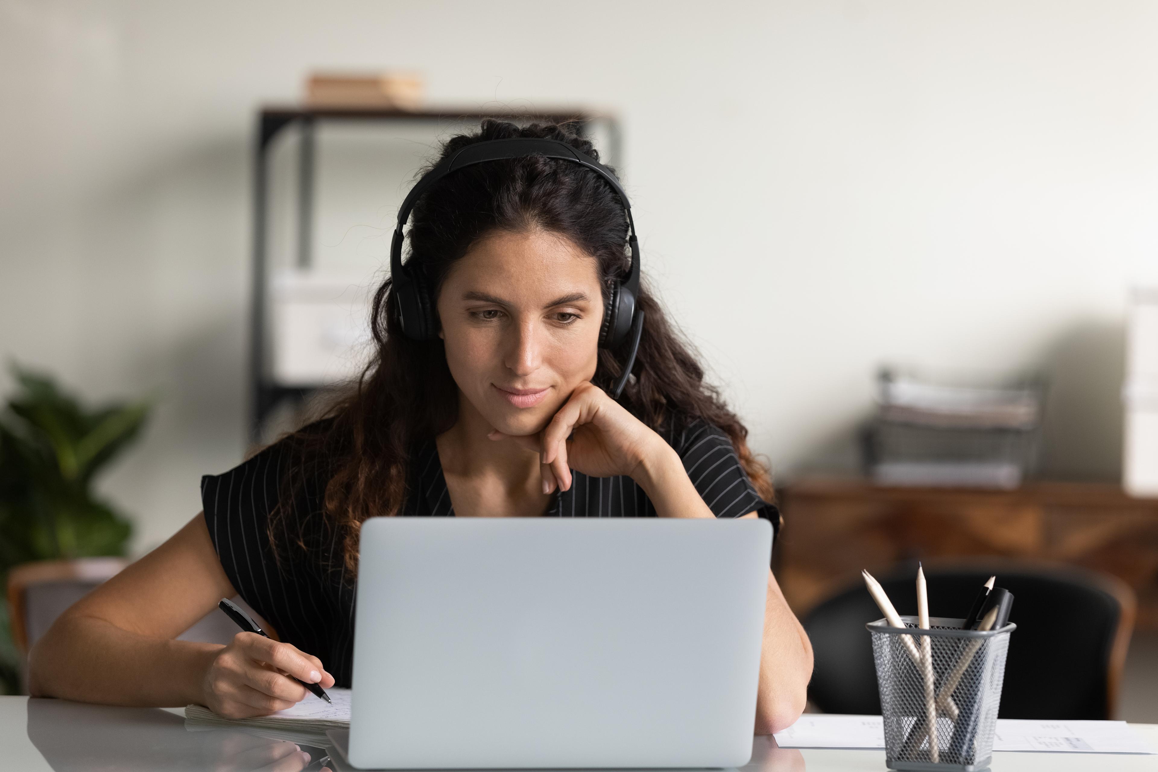 a woman with headphones focused on her laptop, taking notes while listening and looking at screen