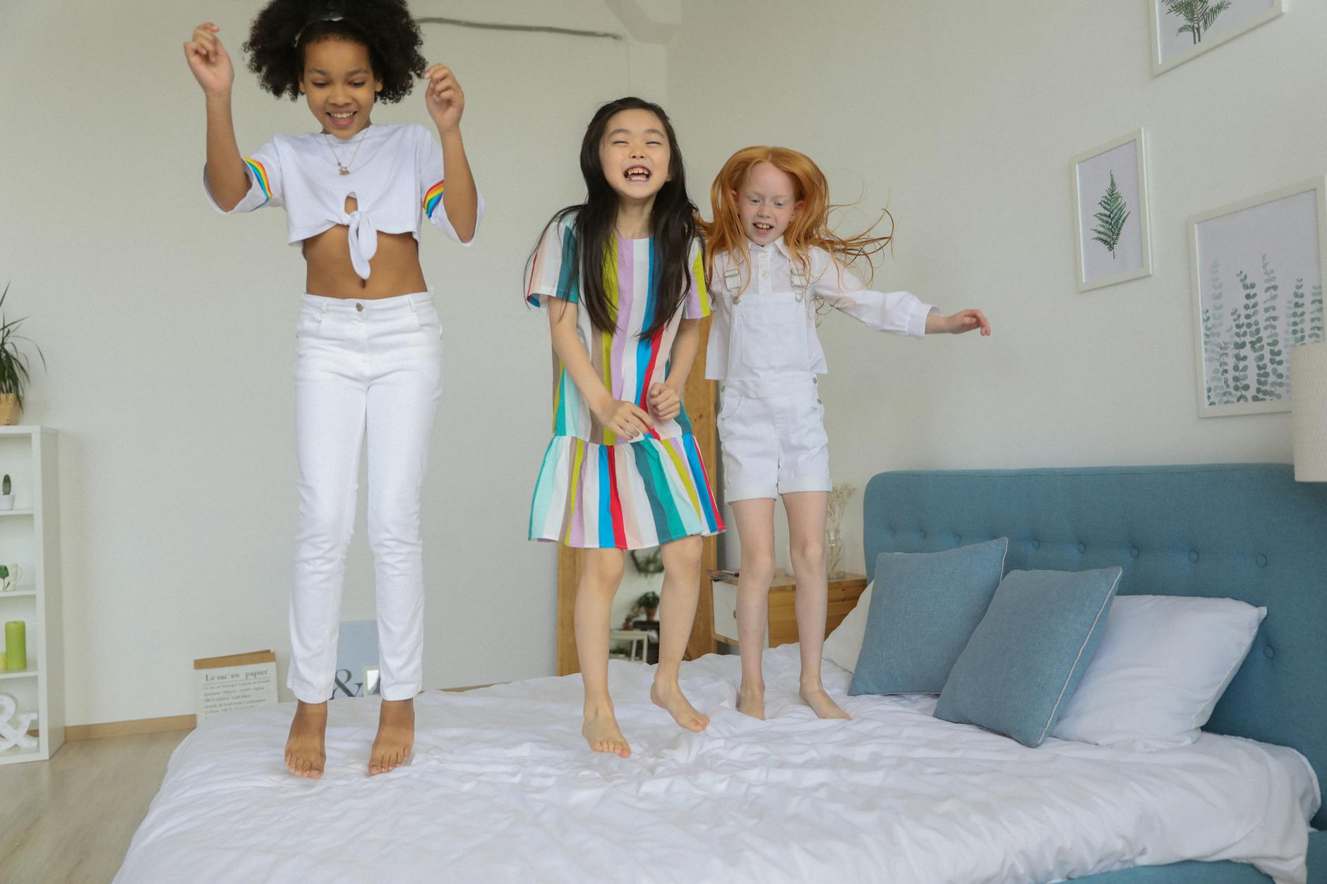 Full body of cheerful multiracial girls laughing and smiling while jumping on bed in modern comfortable room