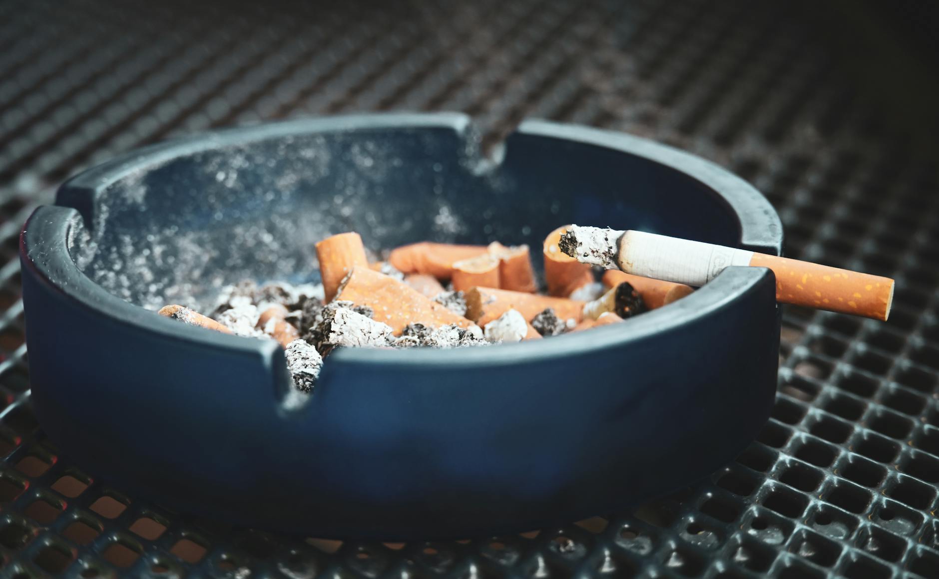 Close-up of cigarette butts and ashes in a blue ashtray with a smoldering cigarette, highlighting smoking habits.