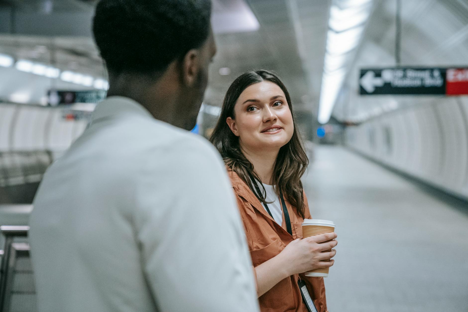 Two adults talking at a subway station platform, holding a coffee cup, during the day.