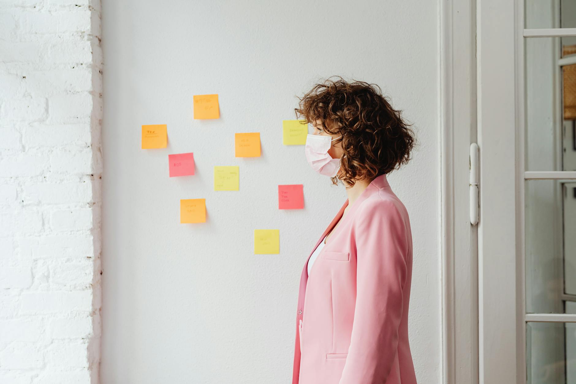 Side view of a businesswoman in a pink blazer with curly hair wearing a face mask indoors.