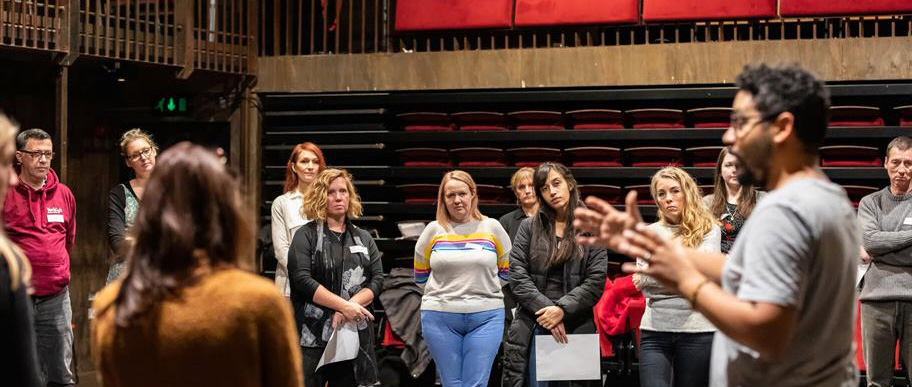 A group of teachers in a circle in a theatre space, absorbed by a man presenting.