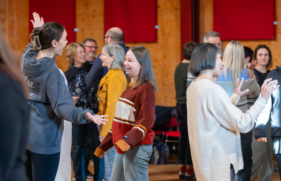 A varied group of teachers, paired off in a large rehearsal room, talking animatedly. 