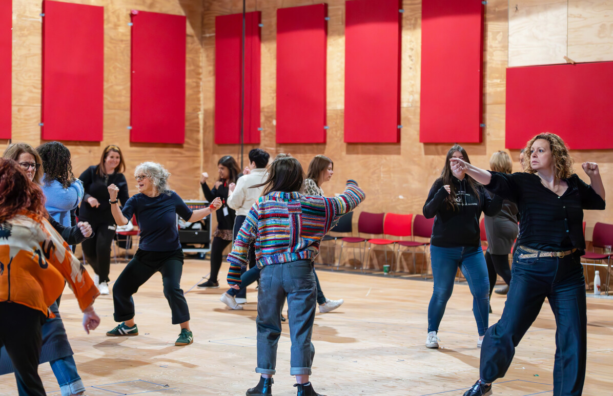 Teachers spread out, in various lively fighting poses in a large, bright rehearsal room. 