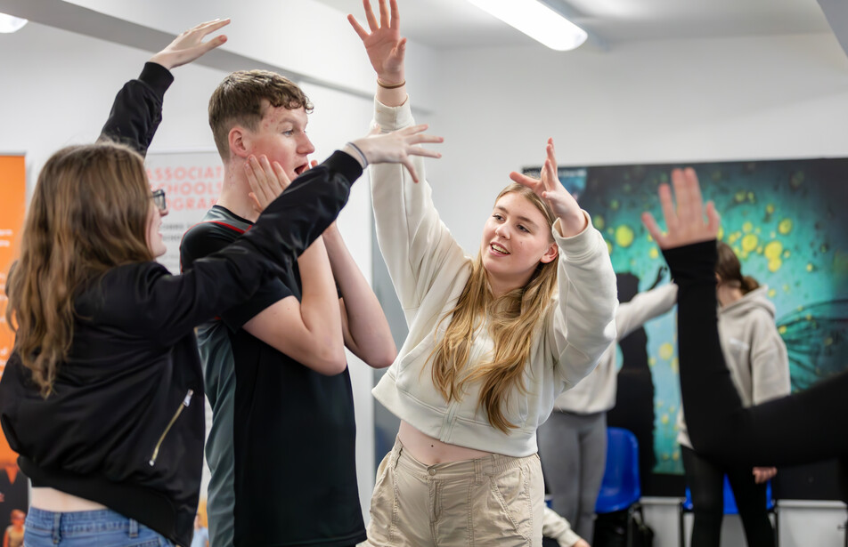 Three older students create a frozen image with their arms in the air in an bright workshop space.