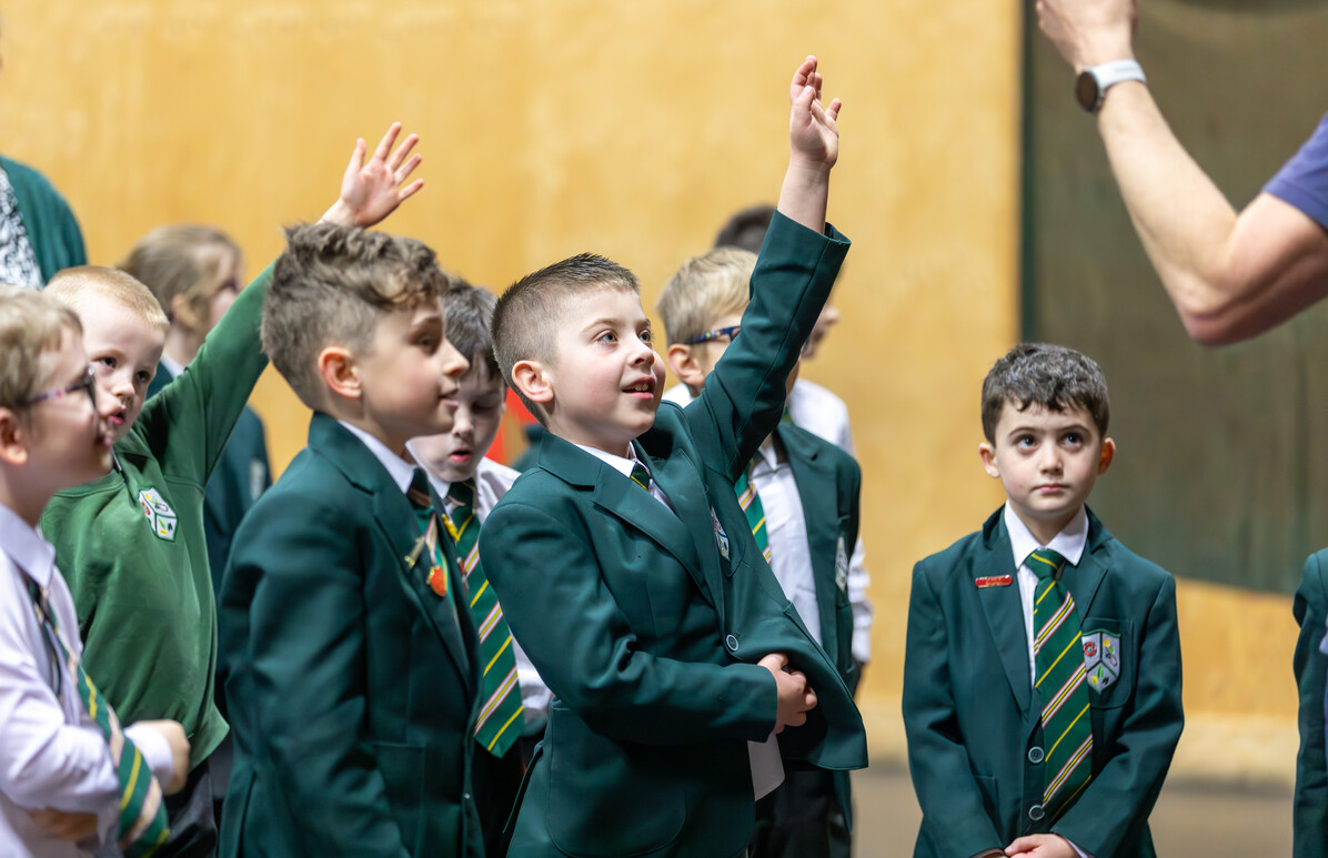 A group of young school children in green blazers and striped ties eagerly raise their hands.