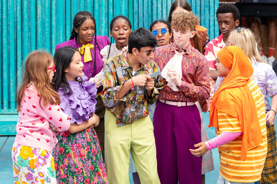 Colourfully dressed Next Generation Act Company members excitedly gather round a messenger on stage.