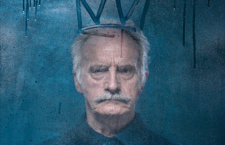 A man in a dark shirt looks at the camera through a wet pane of glass. Water rolls down the glass. A crown has been drawn above his head,