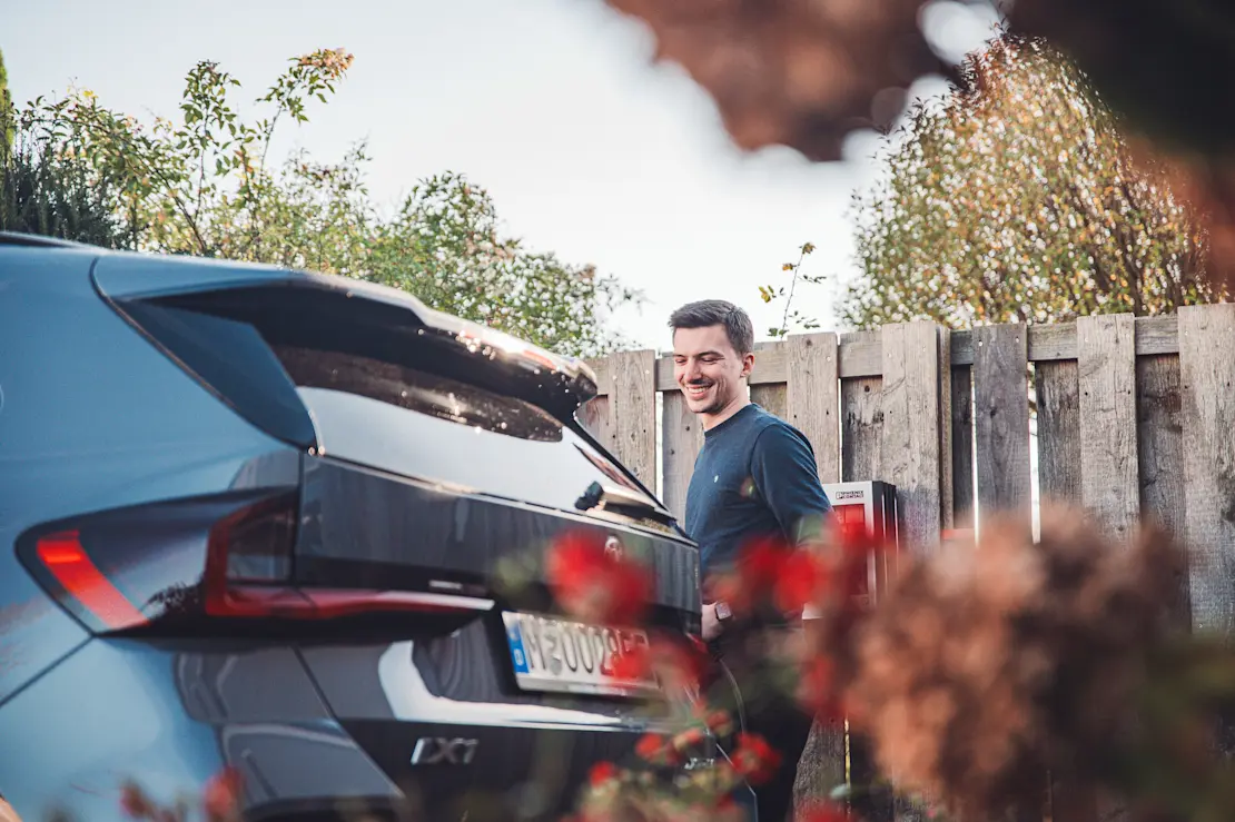 A person stands next to the rear of a grey car with a European license plate. The background features a wooden fence and greenery, with blurred flowers in the foreground.