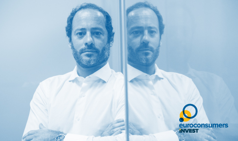 The image features a professional-looking man with short, curly dark hair and a beard, wearing a white button-up shirt. He stands with his arms crossed in an office environment with glass walls and a modern design. On the left side of the image, there is a logo for "Euroconsumers INVEST," incorporating blue and yellow circular elements. The overall composition suggests a corporate and financial investment theme.