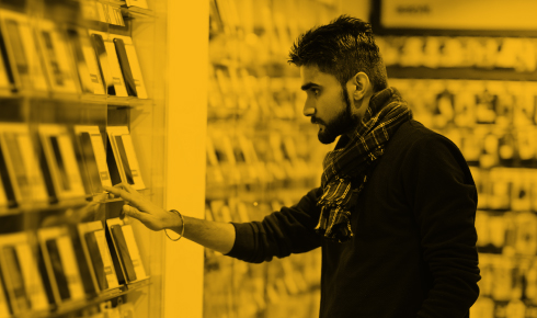 A young man with a beard and dark hair, wearing a black sweater and a checkered scarf, is browsing a selection of smartphones displayed on transparent shelves in a store. He is reaching out to touch or pick up one of the devices, with a focused and contemplative expression. The background consists of more shelves filled with smartphones, creating a sense of abundance and choice. The entire image has a yellow monochromatic overlay.