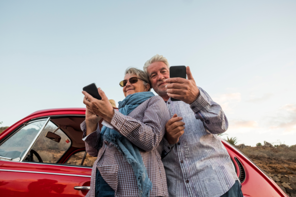 A mature couple leaning against a classic car while they're both on their phones