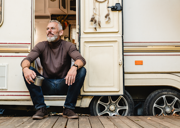 Mature man sitting in the doorway of an RV, looking outside with a mug in hand