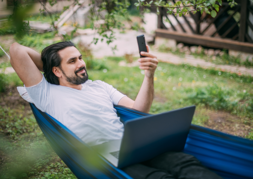 A young man laying in a hammock, looking at his phone while on a laptop