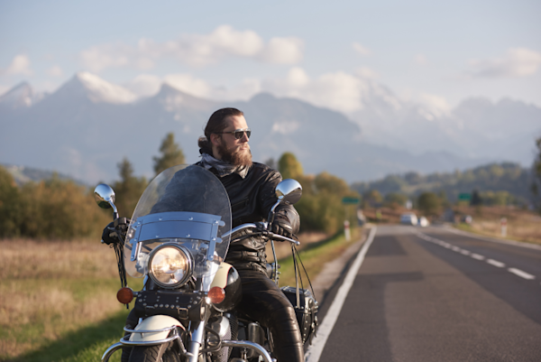 A young man on a motorcycle, looking into the distance with mountain scenery behind him