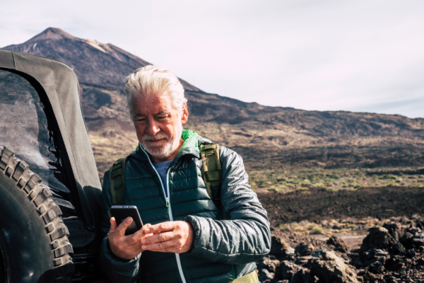 Mature man leaning against a jeep, looking at his cellphone with a mountain behind him