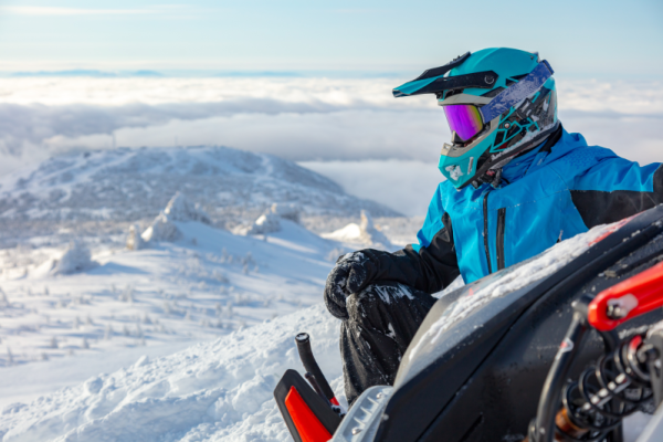 A person in full snowmobile attire looking out at snowy mountains with their snowmobile beside them