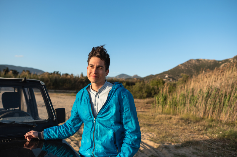 Young man with hand on car hood, looking forward with grassy hills behind him