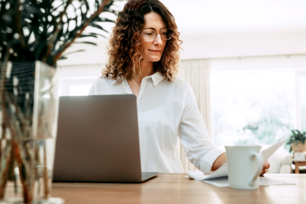 Woman sitting at a table on laptop, looking at a piece of paper