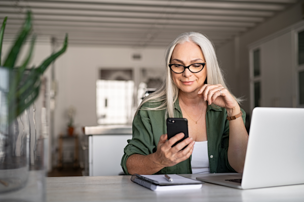 Smiling woman using mobile phone while working at home with her laptop
