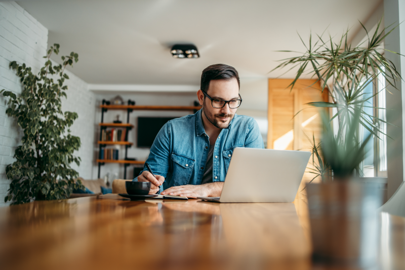 Man taking notes and looking at his laptop while in his home office