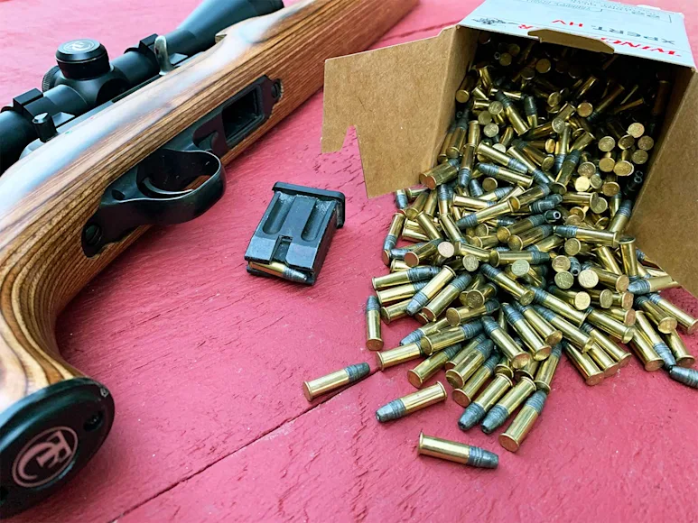 A rifle on a table next to a box of rifle ammunition spilling out onto the table.