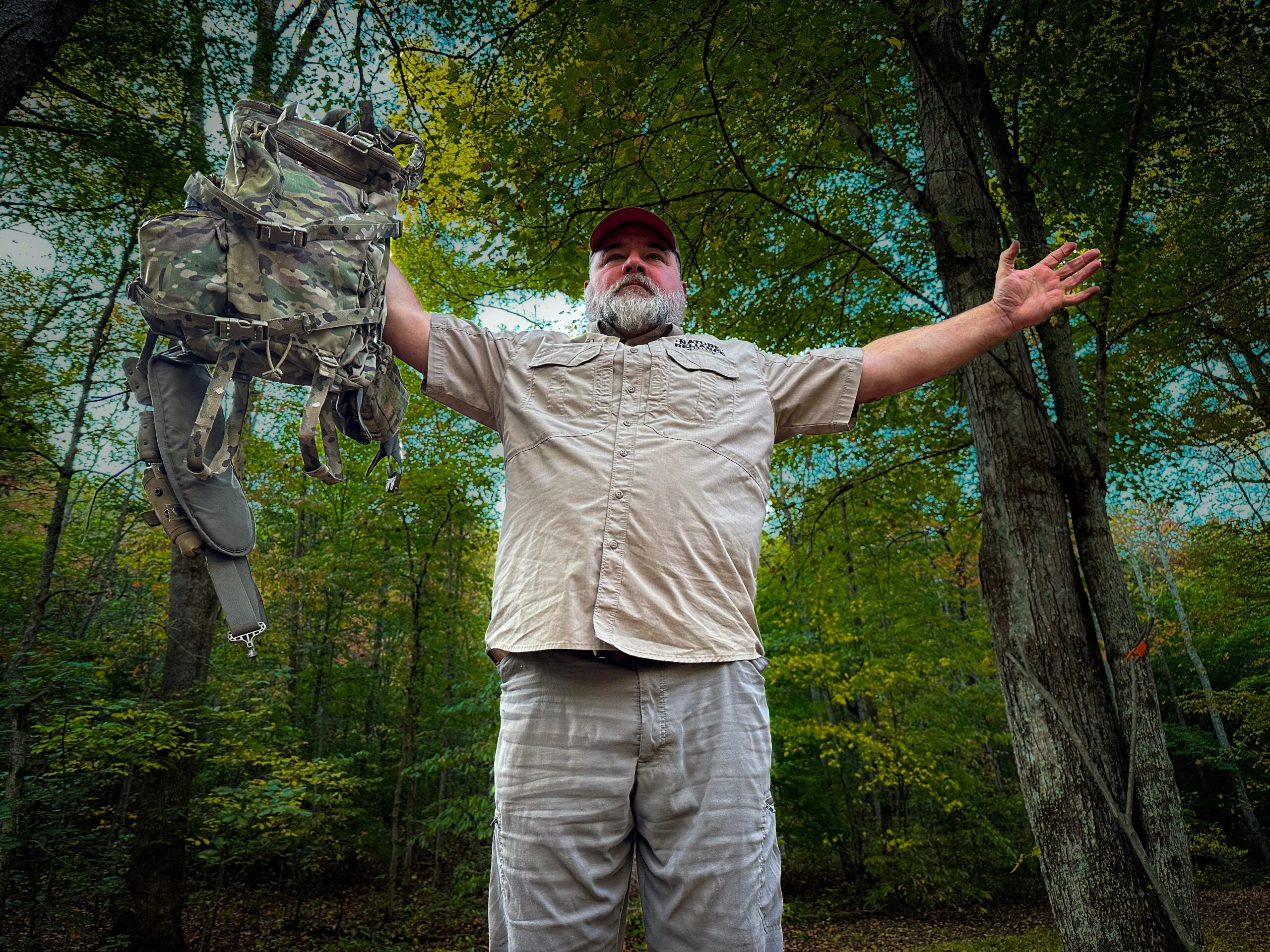 A man in a tan shirt stands in the woods with his arms stretched out