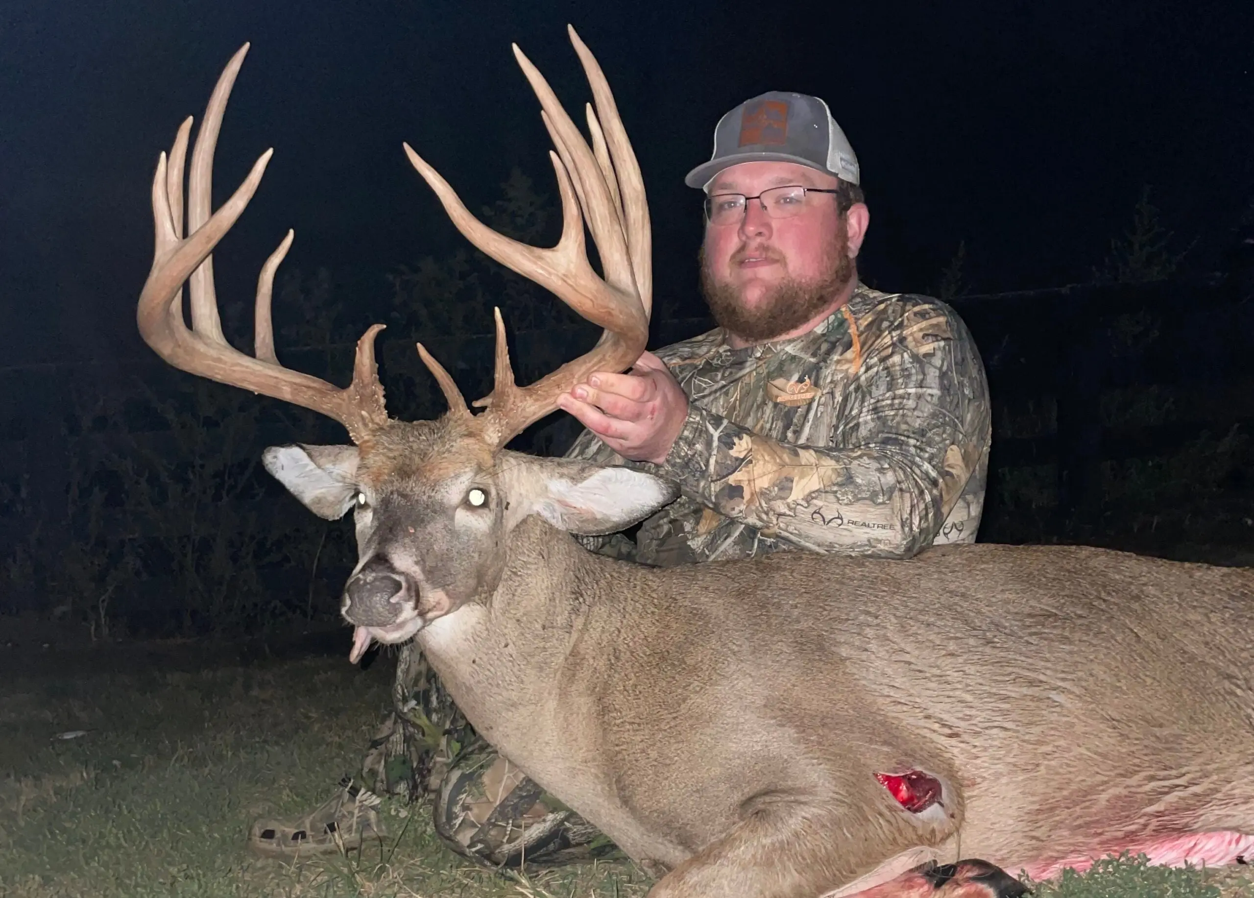 A hunter dressed in camo sits on the ground and shows off a huge whitetail buck.